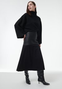 Suéter de punto negro de gran tamaño con cuello alto, combinado con una falda negra ajustada de textura acanalada. Completo con botas de tacón alto negras hasta la rodilla.