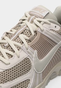 Scarpa Nike Vomero 5 in beige e crema con tessuto in mesh, chiusura con lacci e logo Nike visibile sul lato.
