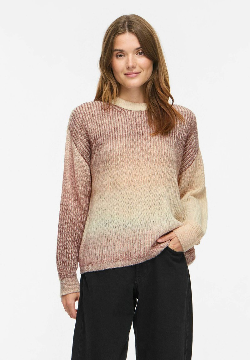 Jeune femme aux cheveux bruns portant un pull tricoté dégradé beige et rose et un pantalon noir, posant devant un fond blanc.
