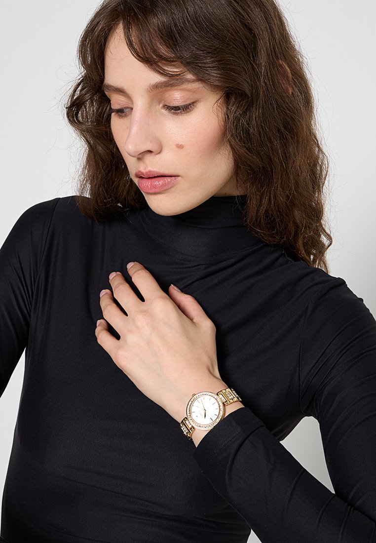 Goudkleurig horloge met een witte wijzerplaat, metalen armbandontwerp, voorzien van duidelijke steentjes en een ronde kast, gedragen op een zwart langemouwen top.