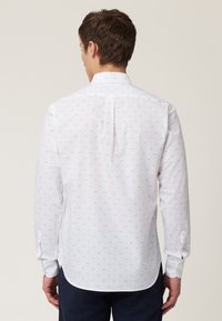 Camisa blanca con un patrón repetido de pequeñas formas en azul claro, de mangas largas, con puños abotonados y un dobladillo recto con pliegue en la parte posterior.