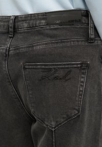 Temno sivi denim hlače z majhnim izvezenim logotipom na zadnjem žepu in elegantno črno nalepko na pasu.