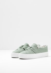Des chaussures slip-on en tissu vert avec un design de nœud à l'avant, une semelle en caoutchouc blanche et une doublure intérieure à pois.