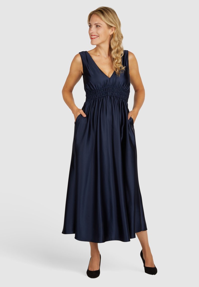 Robe maxi en satin navy avec un décolleté en V, taille smockée et poches latérales ; texture lisse, design sans manches et silhouette fluide.