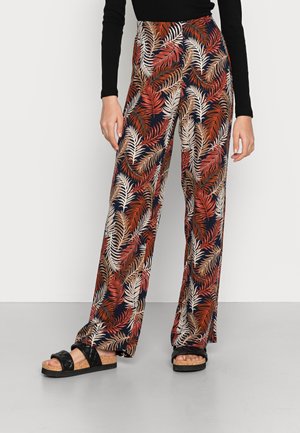 Femme portant un pantalon large à motifs de feuilles rouges, beiges et marron, un haut noir à manches longues et des sandales noires avec semelles en liège.