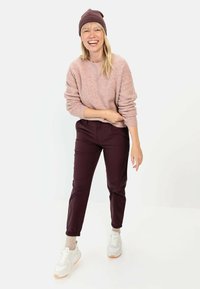 Hellrosa Strickpullover, bordeauxrote tapered Hose und weiße Sneaker mit orangen Akzenten. Das Model trägt eine passende bordeauxrote Mütze.