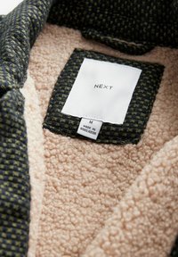 Kollierte Jacke mit einer dunklen gewebten Außenschicht und grünen Akzenten sowie einem weichen beigen Fleecefutter. Etiketten zeigen Marke und Größe an.