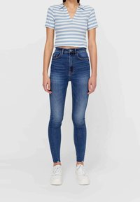 Persona que lleva unos pantalones vaqueros ajustados de talle alto, descoloridos y azul, zapatillas blancas y una camiseta corta de manga corta a rayas azul y blanca.