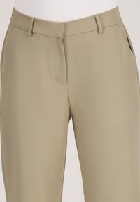 Beige op maat gemaakte broeken met riemlussen, zijzakken en een verborgen sluiting aan de voorkant op een witte mannequin torso.