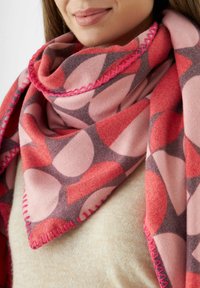 Une femme portant une écharpe à motifs rose, rouge et violet avec des bords cousus sur un pull beige, montrant ses lèvres et son menton.