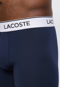 Κοντινή λήψη της μέσης ενός ατόμου που φορά μπλε σκούρα βερμούδα μποξεράκια με λευκή ζώνη μέσης που φέρει το όνομα της μάρκας "LACOSTE" με μαύρα γράμματα.