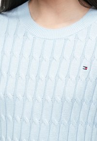 Jasnoniebieski sweter z dzianiny z wzorem w warkocze, okrągłym dekoltem i małym emblematem logo w czerwonym, białym i niebieskim kolorze po lewej stronie klatki piersiowej.