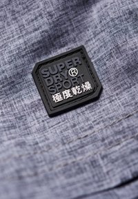 Šedá texturovaná tkanina s černým obdélníkovým nášivkem, na kterém je vyraženo "SUPER DRY SPORT" a pod ním čínské znaky.