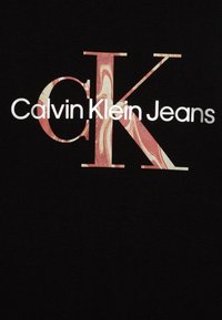 Camiseta negra con un gran logo "K" en tonos rojo y crema veteados, y texto blanco que dice "Calvin Klein Jeans" debajo. Textura de tela suave.