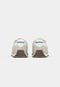 Bežīgi zamšādi sporta apavi ar baltajiem akcentiem, brūnu gumijas zoli un zilo "Reebok" logotipu uz papēža. Aizmugures skats uzsver slāņveida dizainu.