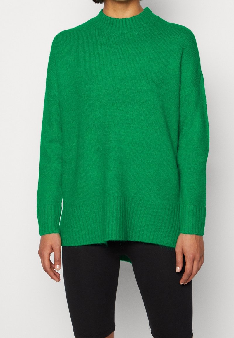Personne portant un pull en maille vert vif à manches longues avec des poignets côtelés, associé à un pantalon noir mi-genou.