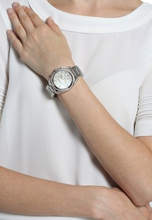 Personne portant un haut à manches courtes blanc avec une montre-bracelet argentée au poignet gauche, la main droite reposant sur la poitrine.