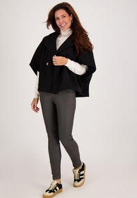 Zwarte poncho met een grote kraag, voorzien van een gouden knop. Gecombineerd met grijze broek en zwarte sneakers met witte strepen.