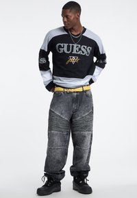 Maglietta a maniche lunghe nera e grigia con logo "GUESS", abbinata a jeans grigi larghi con strisce strutturate e cintura gialla. Stivali neri.