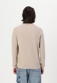 Persoon met schouderlang donkerhaar draagt een beige trui en blauwe denim broek, staande met de rug naar de camera tegen een plain witte achtergrond.