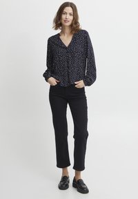 Schwarze Bluse mit langen Ärmeln und V-Ausschnitt, versehen mit einem weißen Pünktchenmuster. Kombiniert mit schwarzen geraden Hosen und schwarzen Slippern.