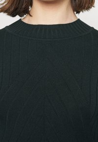 Gros plan d'une personne portant un haut en maille côtelée sombre avec un motif en zigzag et un col rond haut, montrant le cou et une partie des cheveux.