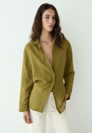 Donna con capelli lunghi e mossi indossa una camicia verde oliva con bottoni abbinata a pantaloni bianchi, in piedi davanti a uno sfondo chiaro e uniforme.