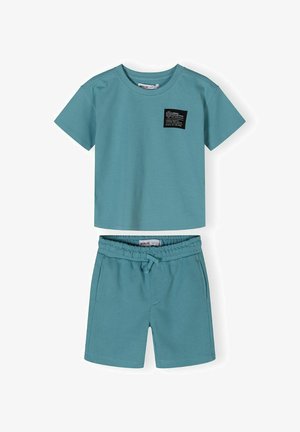 Tirkizna pamučna majica s kratkim rukavima sa crnom zakrpom i odgovarajućim kratkim hlačama. Hlače imaju elastičnu traku za struk i vezicu za podešavanje.