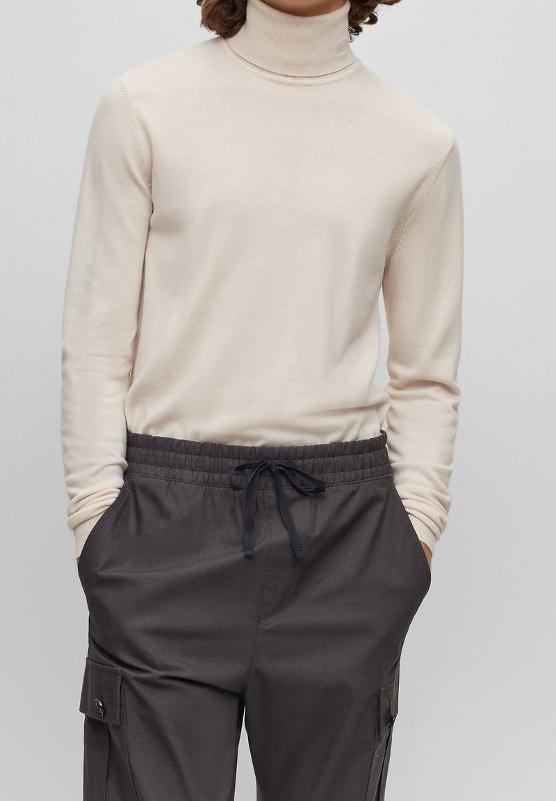 Cremfarbener Rollkragenpullover aus weichem Material, kombiniert mit dunkelgrauen Cargo-Hosen mit elastischem Bund, seitlichen Taschen und Kordelzug.
