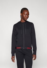 Zwarte rits hoodie van een zachte stof, met een capuchon met trekkoord en een rode "HUGO" logo op de tailleband. Slim fit ontwerp met lange mouwen.