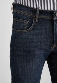 Jeans in denim scuro con una vestibilità slim. Le caratteristiche includono cinque tasche, cuciture a contrasto e una chiusura con bottone e zip visibili.