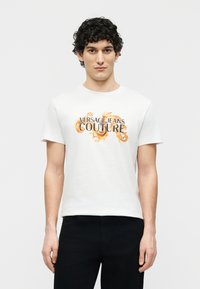 Νεαρός άνδρας με σγουρά μαλλιά, φοράει λευκό t-shirt Versace Jeans Couture με πορτοκαλί διακοσμητικά στροβίλους, συνδυασμένο με μαύρο παντελόνι.