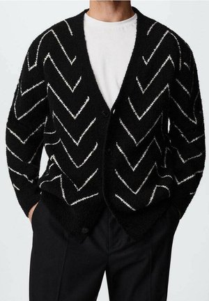 Schwarzer, flauschiger Cardigan mit weißen Zickzackstreifen, getragen über einem weißen Rundhalsausschnittshirt und schwarzen Hosen, die Hände in den Taschen.