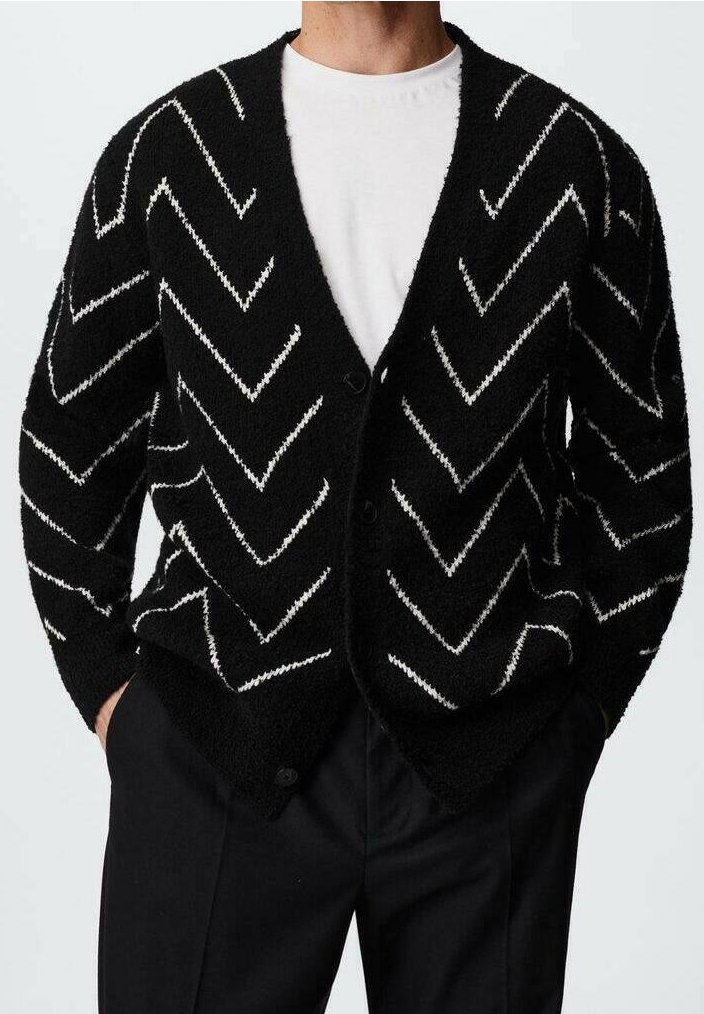 Cardigan noir duveteux à rayures en zigzag blanches, porté par-dessus un t-shirt blanc à col rond et un pantalon noir, les mains dans les poches.