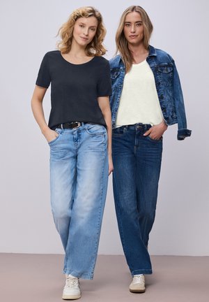 Deux femmes debout côte à côte portant des tenues décontractées : l'une en t-shirt noir et jean large bleu clair, l'autre en haut blanc, veste en jean et jean foncé.