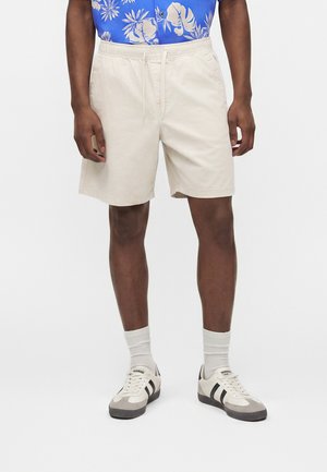 Mężczyzna w beżowych szortach ze sznurkiem, białych skarpetkach i białych trampkach z czarnymi paskami, stojący na jednolitym tle.