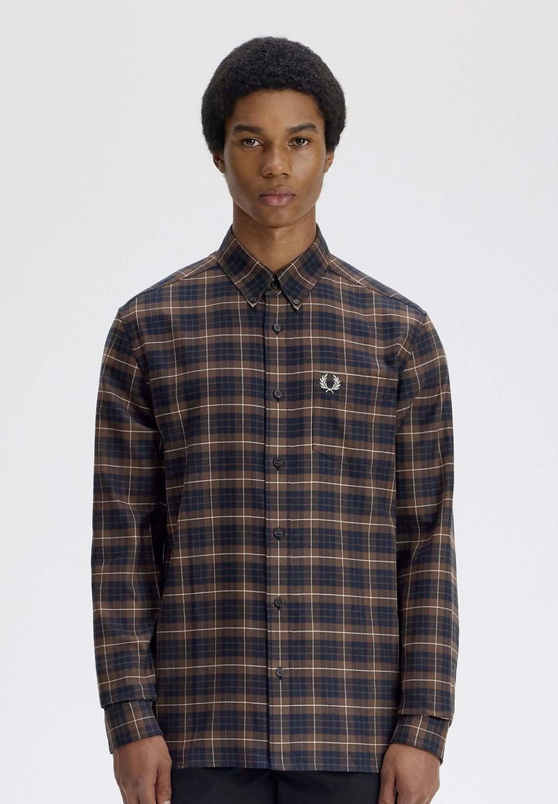 Jeune homme portant une chemise à carreaux marron et bleu marine à boutons avec un petit logo blanc brodé sur la poche poitrine.