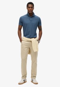 Conjunto masculino que incluye una camiseta polo azul, pantalones beige y un suéter de punto trenzado color crema atado a la cintura. Zapatillas blancas con detalles en negro.