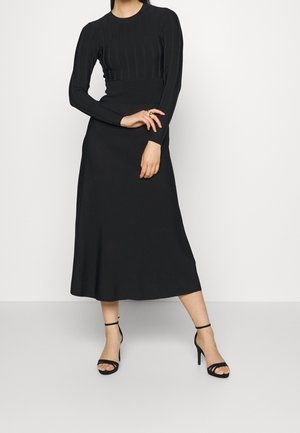 Czarna prążkowana sukienka z długim rękawem, dopasowanym gorsetem i zwiewną midi spódnicą; zestawiona z czarnymi sandałami na obcasie z paskami.