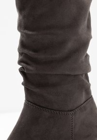 Bota de ante gris hasta la rodilla con textura arrugada y detalles de costura en el tobillo. Acabado suave y tela blanda.