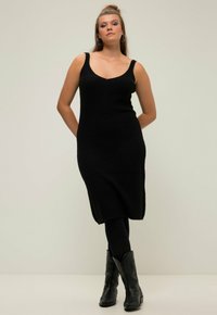 Zwart geribbeld gebreid jurk met een V-hals en brede banden, gecombineerd met zwarte legging en cowboylaarzen, tegen een effen achtergrond.