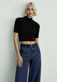Crna kratka turtleneck pulover u rebrastom materijalu, uparen sa širokim tamnoplave traperice. Ima crni remen s metalnom kopčom.
