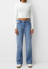 Camiseta blanca de manga larga recortada, vaqueros azul claro de pierna ancha y zapatillas blancas con suelas marrón claro. Los vaqueros tienen un diseño tradicional de 5 bolsillos.