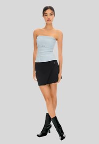 Femme portant un top léger bleu sans manches, une mini-jupe asymétrique noire et des bottes à talons noires, se tenant contre un fond clair.