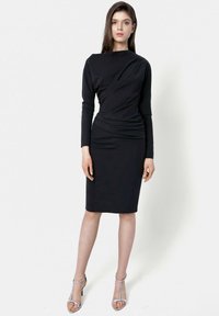 Czarna długa sukienka z długimi rękawami o dopasowanej sylwetce, z drapowaną przodu i długości do kolan. Noszona z srebrnymi szpilkami na paski. Gładka tekstura tkaniny.
