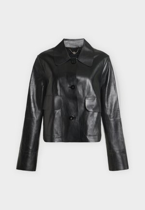 Femme portant une veste en cuir noire et un pantalon, un haut blanc, tenant un sac à main blanc, debout devant un fond clair uni.