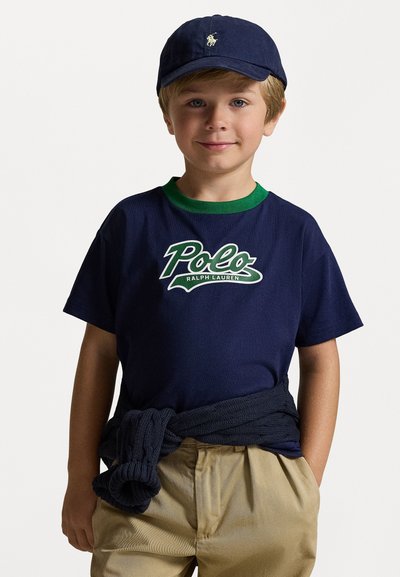 Tamsiai mėlynas medvilninis marškinėlis su žaliu apykakle ir baltu logotipu, derinamas su smėlio spalvos kelnėmis ir tamsiai mėlynu megztiniu. Taip pat yra tamsiai mėlyna kepurė.