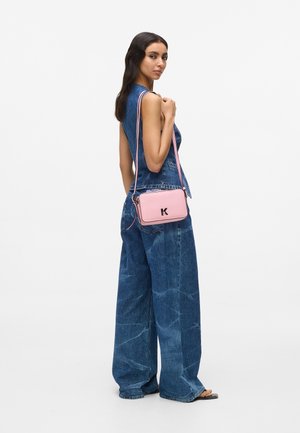 Roze crossbodytas met zwart logo, rechthoekige vorm, glad leren textuur en verstelbare riem, gedragen over een spijkerbroekoutfit.