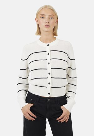 Młoda kobieta o krótkich blond włosach, ubrana w biały kardigan zapinany na guziki z czarnymi poziomymi paskami i czarne spodnie, z rękami w kieszeniach.