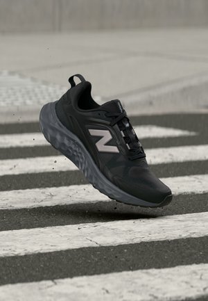 Черна маратонка New Balance във въздуха над пешеходна пътека на асфалтов път с малки частици гравий.